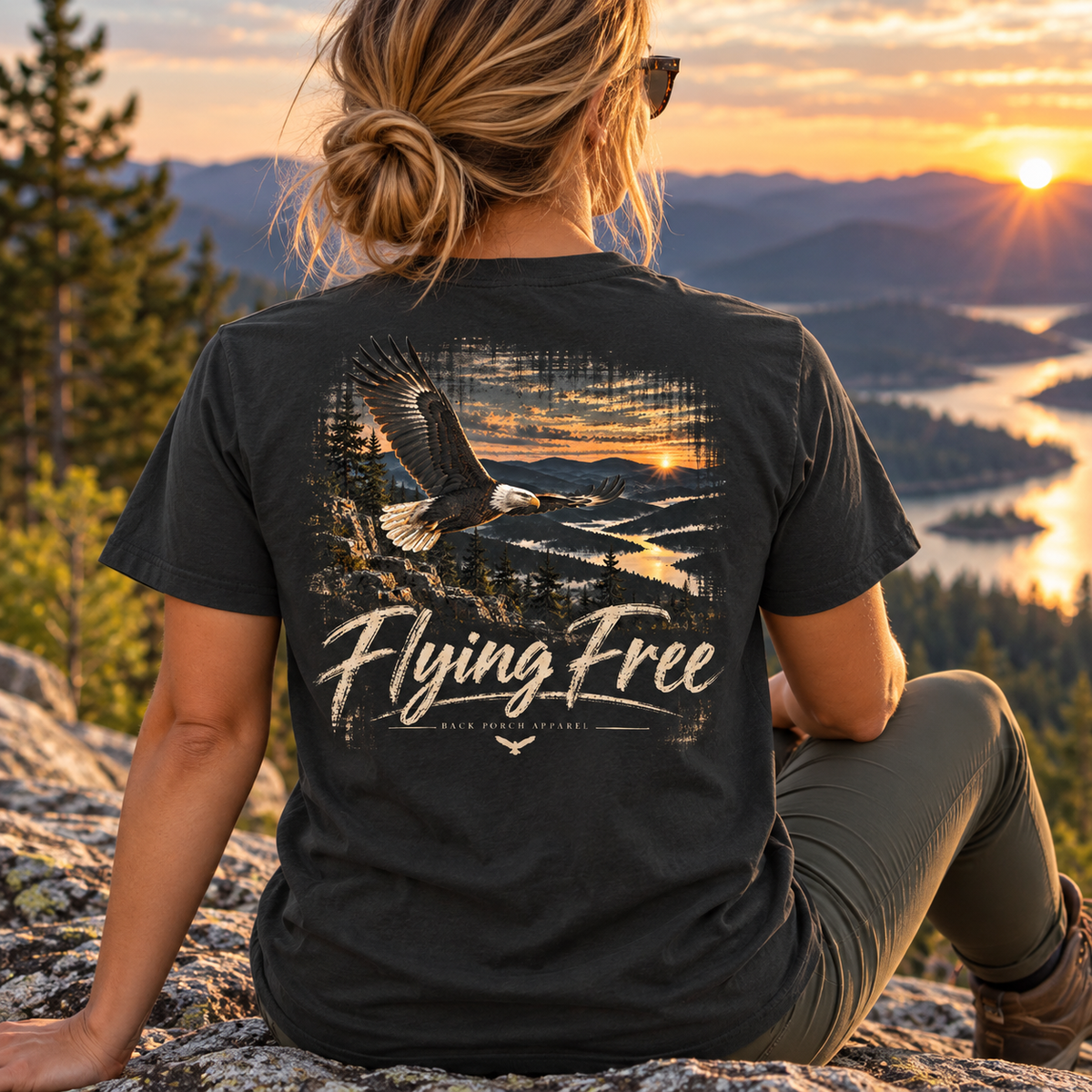 A woman sits on a rocky mountain overlook at sunset wearing a black graphic t-shirt. The back of the shirt features a large graphic of a bald eagle flying over a lake and forest, with the text 'Flying Free' and 'Bald Eagle National Park' printed below.