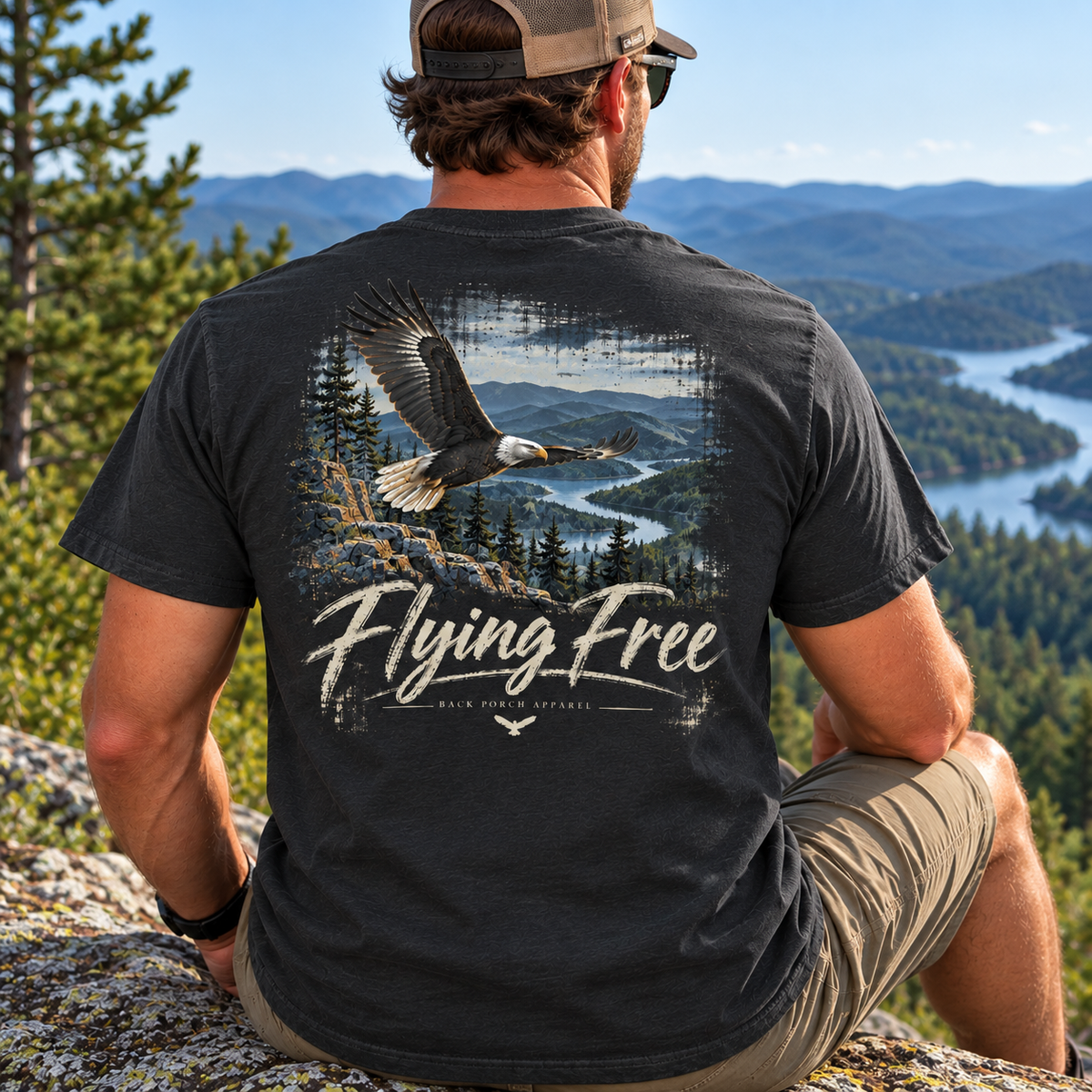 A man sitting outdoors on a rock overlooking a mountain lake landscape, shown from the back wearing a dark charcoal t-shirt with a large graphic of a flying bald eagle and the text 'Flying Free'.