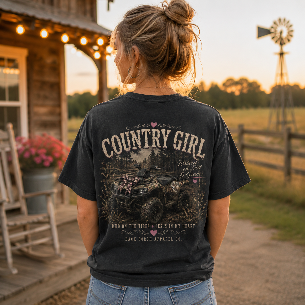A woman stands outdoors at sunset wearing a black graphic T-shirt with a large back print reading "COUNTRY GIRL" and an ATV scene, paired with blue jeans. A rustic cabin, porch lights, fence, and windmill are visible in the background.