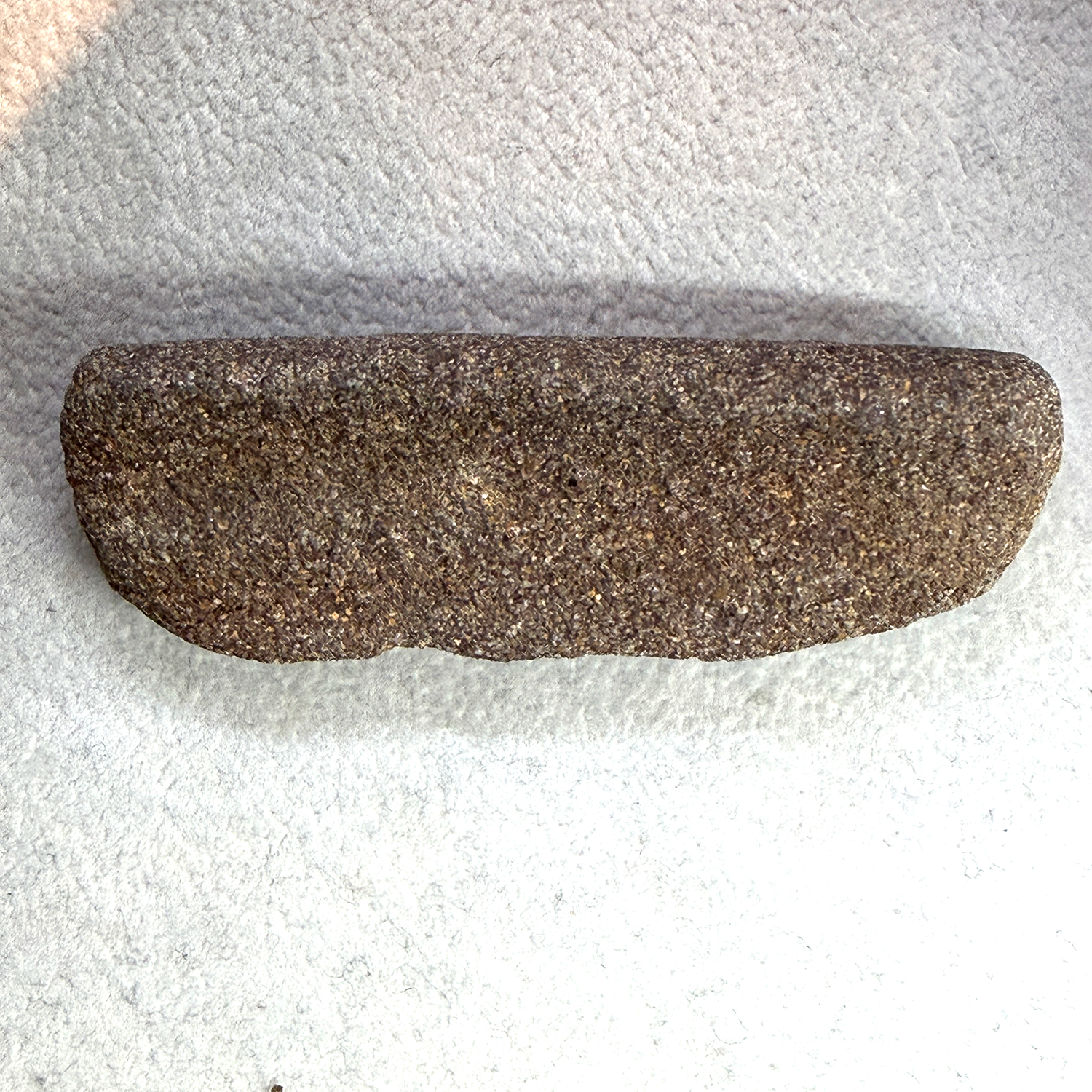 A small, irregularly shaped dark brown stone or rock sample with a rough, speckled texture, photographed on a white textured fabric background.