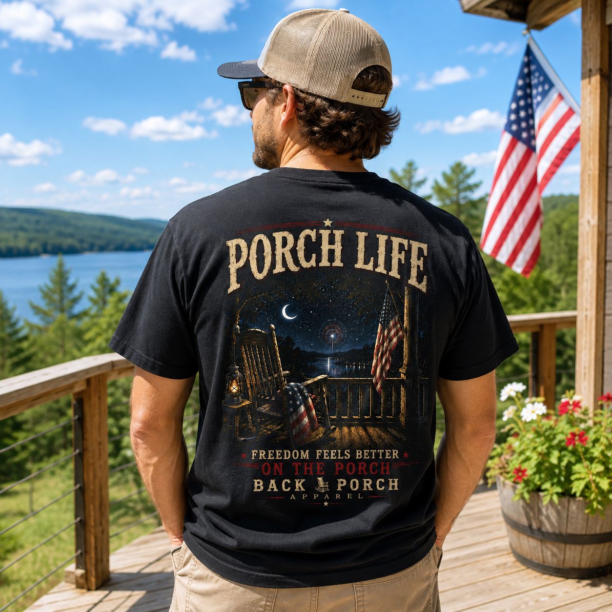A man stands on a wooden porch with a scenic lake and forest background, wearing a black graphic t-shirt with 'PORCH LIFE' printed on the back. The shirt features a detailed graphic of an outdoor rocking chair scene at night with American flags and the text 'FREEDOM FEELS BETTER ON THE PORCH BACK PORCH APPAREL'.
