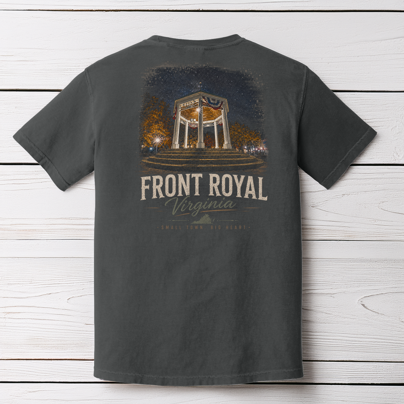 A dark charcoal gray t-shirt shown from the back, featuring a graphic print of the Front Royal Veterans Memorial Garden in Virginia. The graphic includes a night scene of the monument with warm lights and the text 'FRONT ROYAL Virginia' in a distressed cream font, laid flat on a white wood plank background.