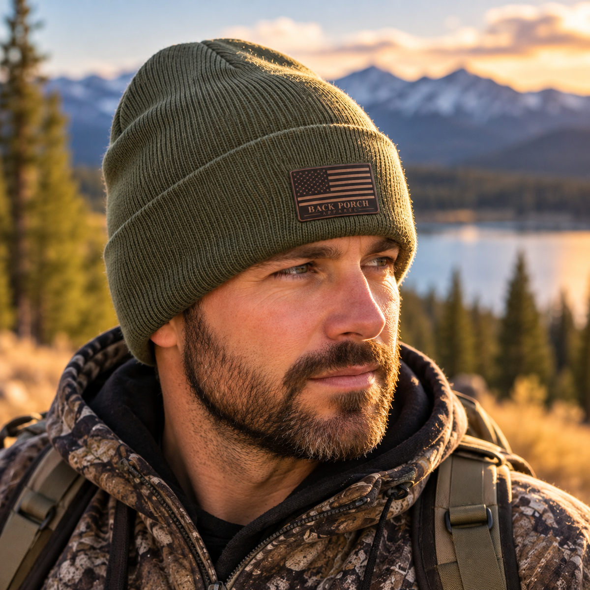 Man wearing an olive drab green knit beanie with a small dark brown leather American flag patch by Back Porch Apparel, outdoors in a mountain setting.