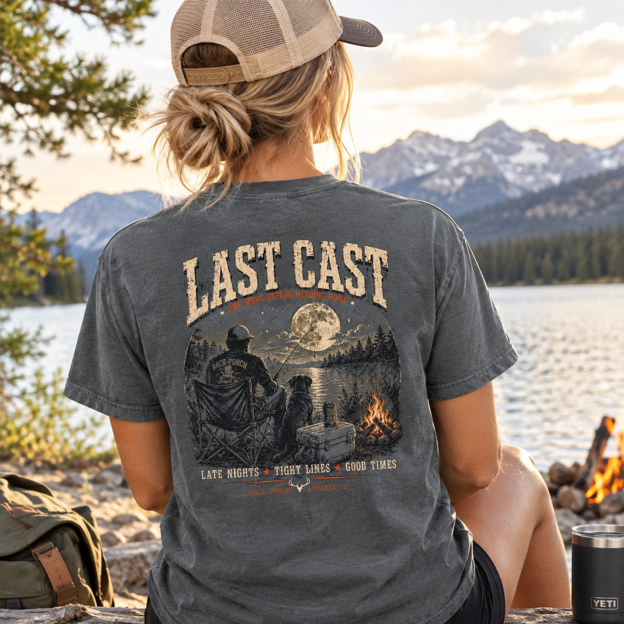 A woman seen from behind wearing a dark gray graphic t-shirt with a campsite scene on the back, sitting by a lake with mountains in the background.