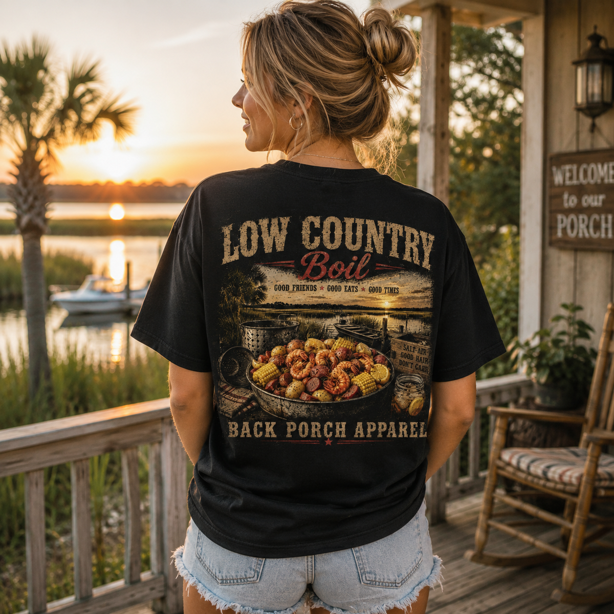 A woman stands on a wooden porch at sunset wearing a black graphic T-shirt with a large back print reading 'LOW COUNTRY Boil' and 'BACK PORCH APPAREL,' showing a pot of shrimp and grits. The background includes water, palm trees, a rocking chair, and a porch sign.