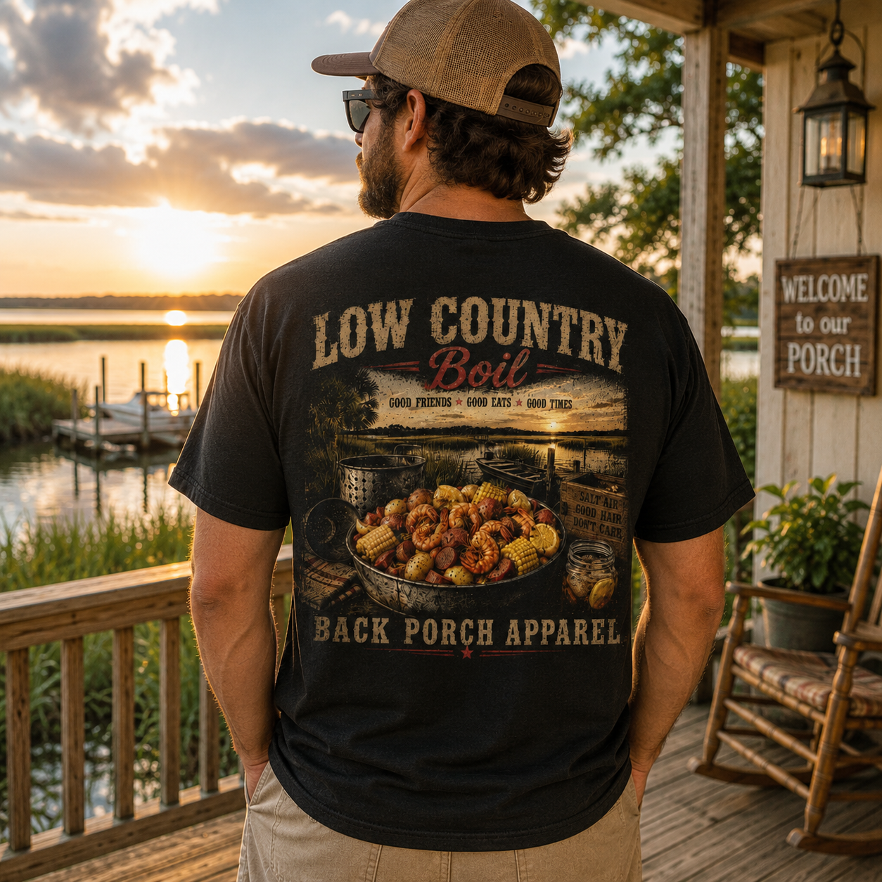 A man stands on a wooden porch at sunset wearing a black graphic T-shirt from the back. The shirt features a large printed design with the text 'LOW COUNTRY Boil' and 'BACK PORCH APPAREL' over an autumnal pond scene with a pot of corn and boils.