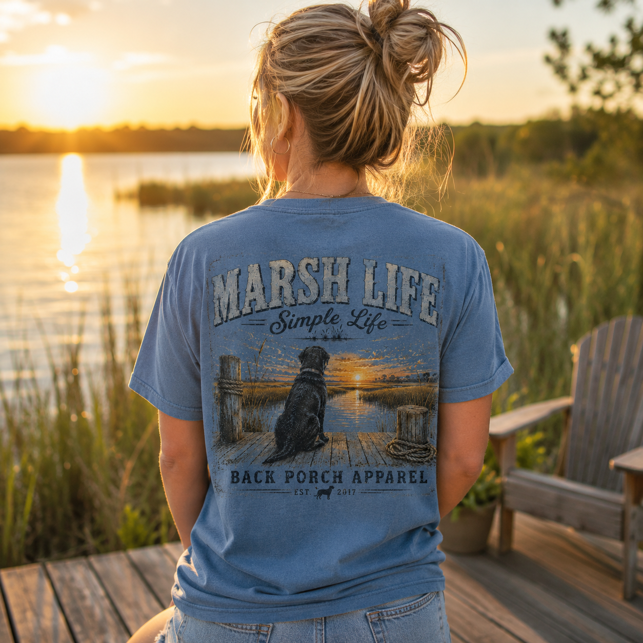A woman stands on a wooden dock by a lake at sunset wearing a blue graphic T-shirt. The back of the shirt features a large printed design with the text 'MARSH LIFE Simple Life BACK PORCH APPAREL' and an illustration of a dog sitting on a dock at the water's edge.