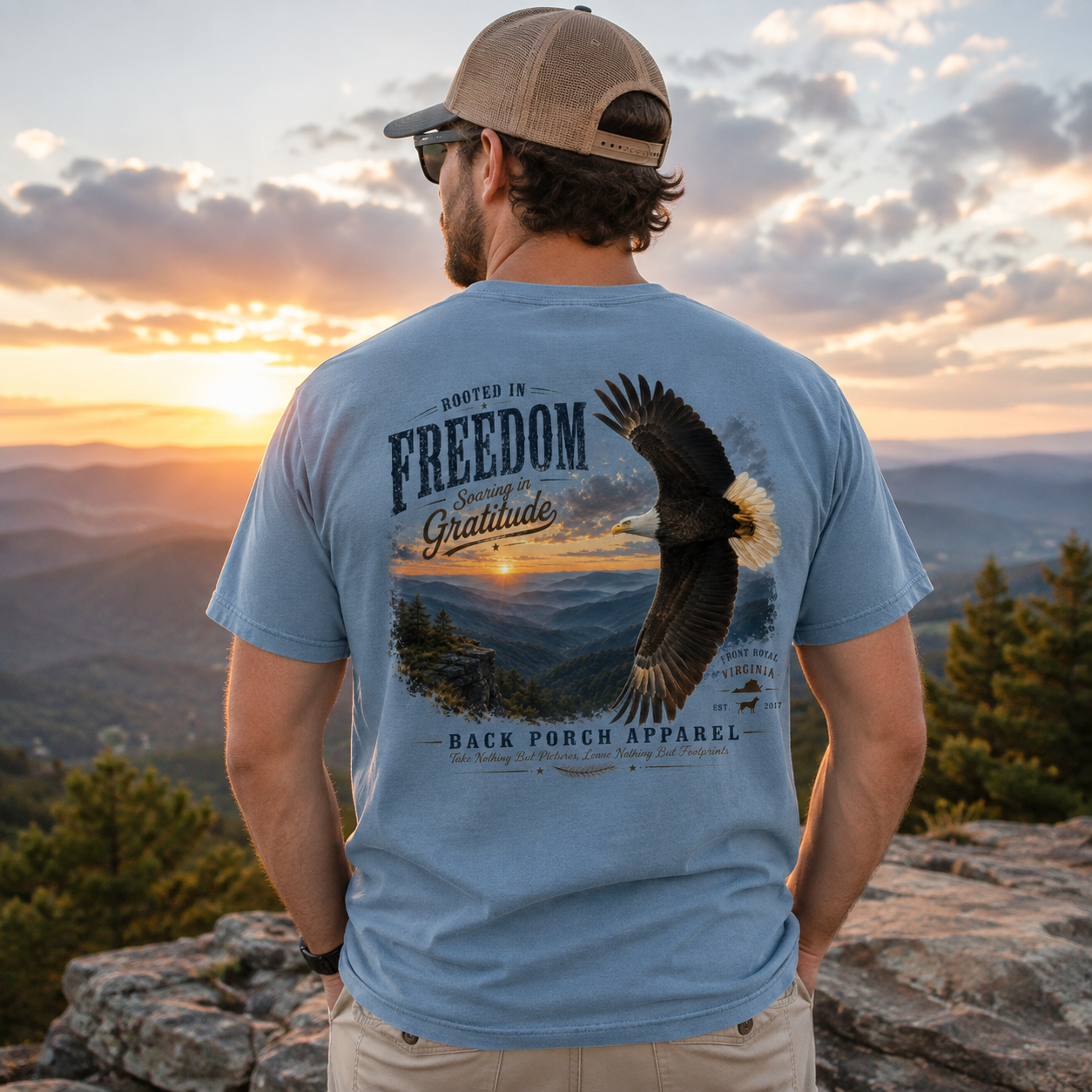 A man wearing a light blue t-shirt with a large eagle and mountain graphic on the back, photographed from behind against a sunset mountain landscape. The t-shirt features the text 'ROOTED IN FREEDOM Gratitude Back Porch Apparel'.