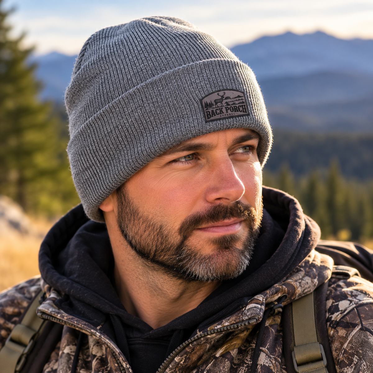 Man wearing a Heather Grey cuffed knit beanie with a small charcoal leather Back Porch Apparel whitetail deer and mountain patch, dressed in camouflage outdoor clothing with a mountain landscape in the background.