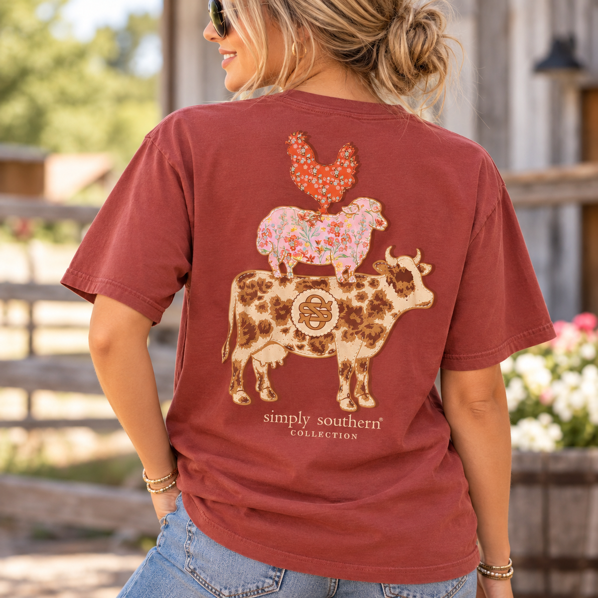 Woman wearing a maple-colored Simply Southern Farm Animals T-shirt with a floral cow, pink sheep, and patterned chicken graphic on the back in a rustic outdoor setting.