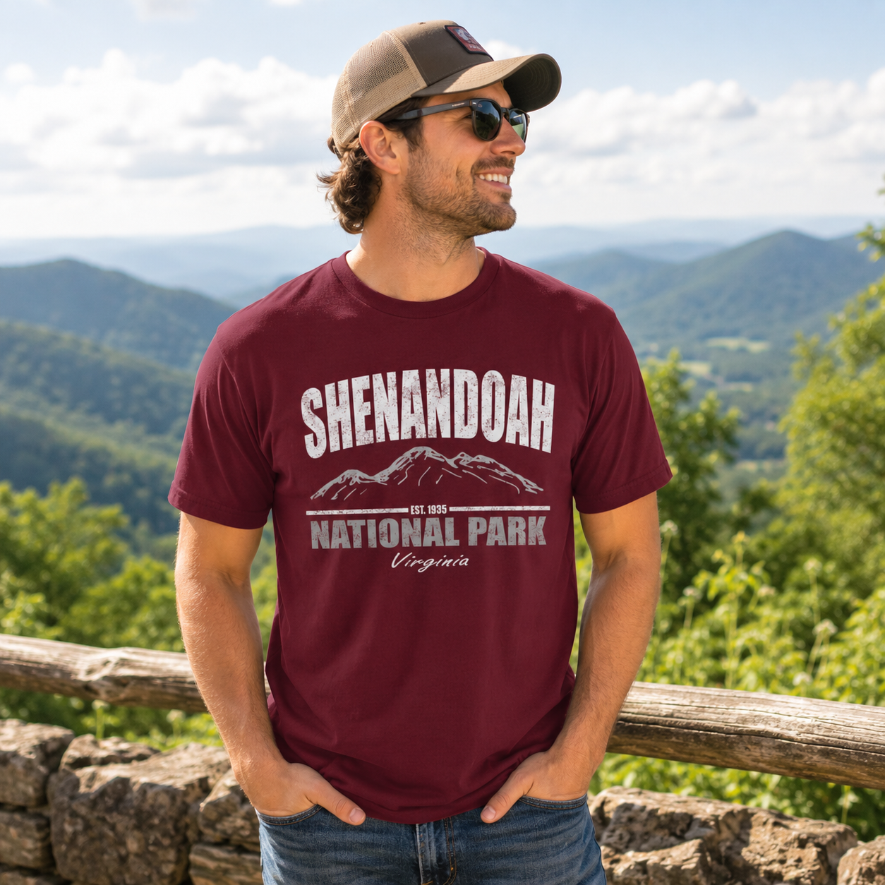 A man wearing a maroon t-shirt with a white graphic that reads 'SHENANDOAH NATIONAL PARK Virginia' and a tan baseball cap, standing outdoors in front of a scenic mountain landscape.