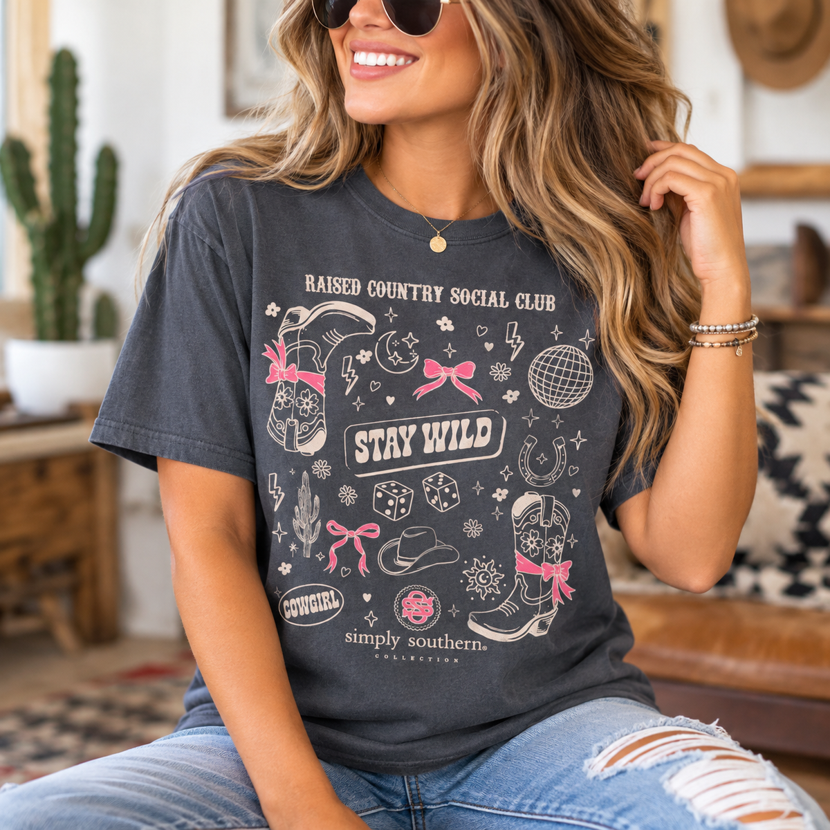 Woman wearing a Simply Southern premium mineral washed Graphite T-shirt with Raised Country Social Club Stay Wild Western cowgirl graphic on the front, styled with distressed denim jeans.