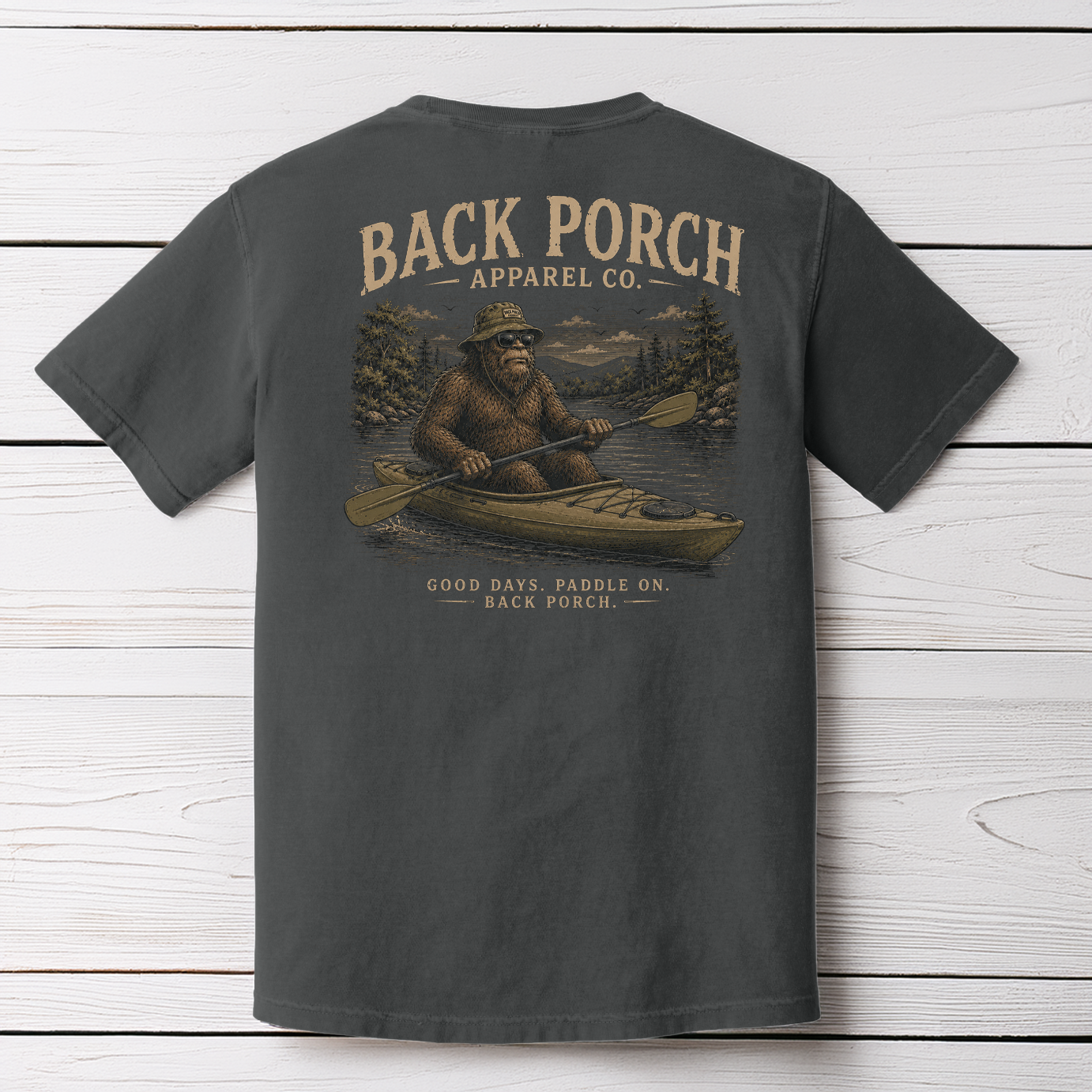 Bigfoot kayaking graphic t-shirt on Comfort Colors garment-dyed tee featuring Sasquatch paddling a kayak through a mountain river with Back Porch Apparel branding