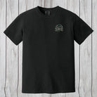A black short-sleeve crewneck t-shirt with a small camouflage-patterned circular chest graphic, displayed flat against a light gray wood plank background.