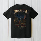 Black Comfort Colors t-shirt featuring patriotic porch life artwork displayed on a white wood background.