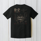 Black Comfort Colors B-2 Spirit stealth bomber t-shirt displayed on a white wood background.