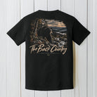 A black short-sleeve t-shirt with a large back graphic showing a bear in a mountain landscape and the text 'The Back Country' and 'BACK POUND APPAREL', displayed against a white wood plank background.