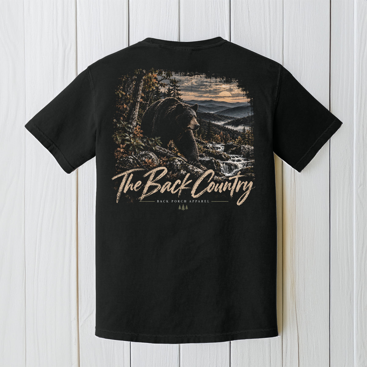 A black short-sleeve t-shirt with a large back graphic showing a bear in a mountain landscape and the text 'The Back Country' and 'BACK POUND APPAREL', displayed against a white wood plank background.