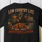 Close-up of the Low Country Life Bigfoot graphic showing a crawfish boil, corn, shrimp, Southern swamp scenery, and the phrase "Boil First, Questions Later" on a Black Comfort Colors t-shirt.