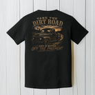 Black Comfort Colors Bigfoot truck t-shirt displayed on a white wood background featuring a classic pickup on a country dirt road.