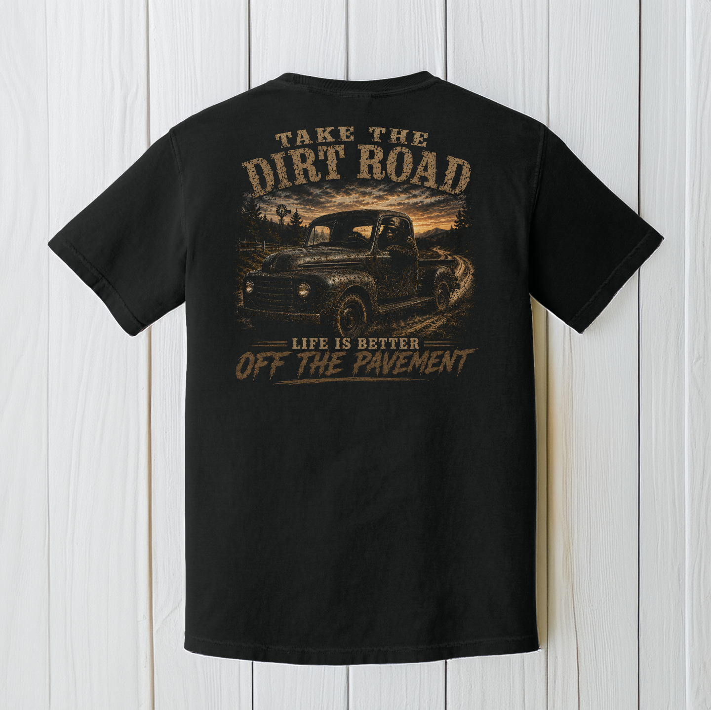 Black Comfort Colors Bigfoot truck t-shirt displayed on a white wood background featuring a classic pickup on a country dirt road.