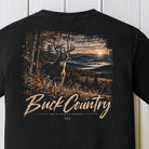 A black short-sleeve t-shirt featuring a large graphic print on the back. The graphic depicts a deer in a forest at sunset with the text 'Buck Country' and 'BACK PORCH APPAREL' underneath, displayed against a white wood-paneled background.