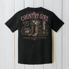 Black Comfort Colors t-shirt hanging on a wood background featuring a rustic country girl western boots and wildflower graphic design.