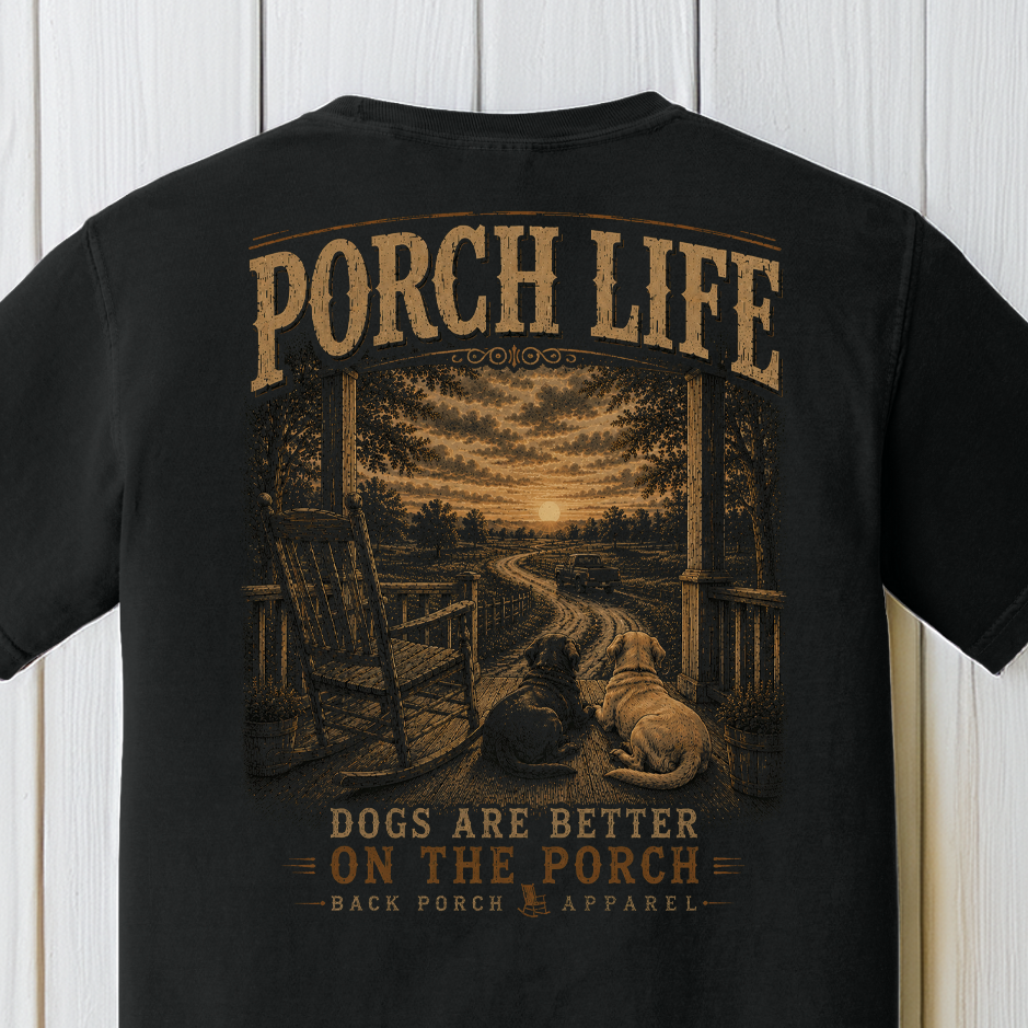 Close-up of the Porch Life Dogs Are Better on the Porch artwork showing two dogs, a rocking chair, winding country road, and sunset on a Black Comfort Colors t-shirt.