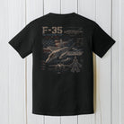 Black Comfort Colors F-35 fighter jet t-shirt displayed on a white wood background.