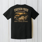 Black Comfort Colors t-shirt displayed on a wood background featuring a southern chicken fried steak graphic with rustic farmhouse porch artwork.
