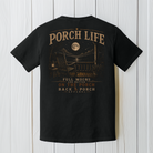 Black Comfort Colors porch life t-shirt displayed on a white wood background featuring a full moon, rocking chair, lantern, and peaceful nighttime country porch scene.