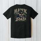 A black short-sleeve t-shirt shown from the back, featuring a large graphic print of a hawk with 'HAWK DAD' written in distressed white and green lettering. The t-shirt is displayed flat against a white wood plank background.