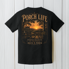 Black Comfort Colors Porch Life graphic t-shirt displayed on a white wood background featuring a peaceful country sunrise viewed from a front porch rocking chair.