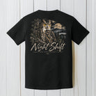 A black short-sleeve t-shirt shown from the back, featuring a large graphic print of a owl in a forest with a moon and the text 'Night Shift' and 'Back Pack Apparel'. The shirt is displayed flat against a white wood plank background.