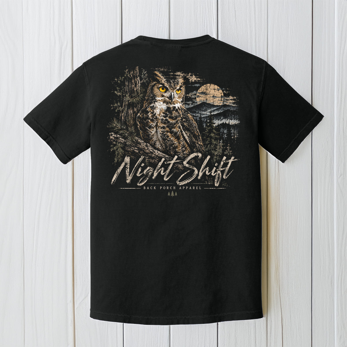 A black short-sleeve t-shirt shown from the back, featuring a large graphic print of a owl in a forest with a moon and the text 'Night Shift' and 'Back Pack Apparel'. The shirt is displayed flat against a white wood plank background.