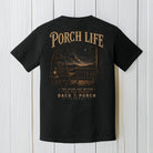 Black Comfort Colors t-shirt featuring a Porch Life nighttime mountain porch scene displayed on a white wood background.