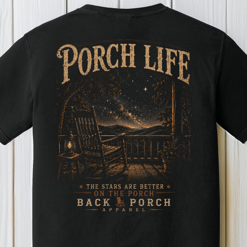 Rustic nighttime porch graphic with rocking chair, lantern, mountains, and star-filled sky on a black Comfort Colors shirt.
