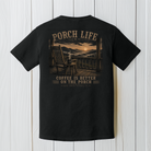 Black Comfort Colors porch life t-shirt displayed on a white wood background featuring a mountain sunrise, rocking chair, and coffee mug with Coffee Is Better on the Porch artwork.