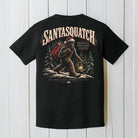 A black short-sleeve graphic T-shirt shown flat against a white wood plank background. The back of the shirt features a large illustrated design of a Bigfoot-like figure wearing a Santa hat and red backpack, with the text 'SANTASQUATCH' and 'BELIEVE IN CHRISTMAS LEGENDS'.