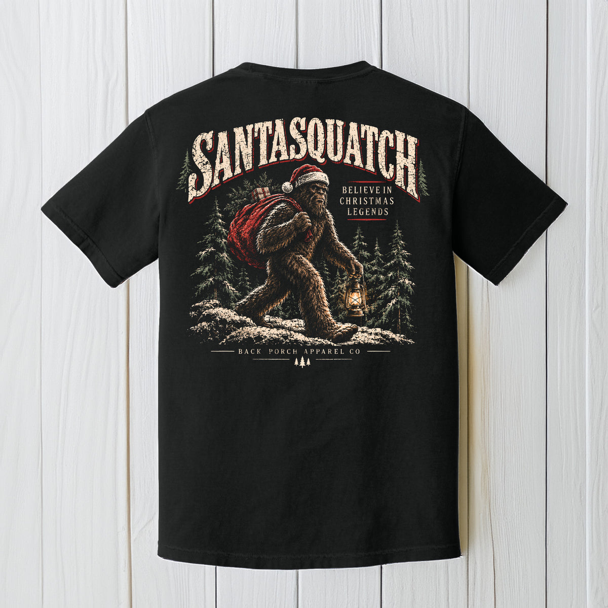 A black short-sleeve graphic T-shirt shown flat against a white wood plank background. The back of the shirt features a large illustrated design of a Bigfoot-like figure wearing a Santa hat and red backpack, with the text 'SANTASQUATCH' and 'BELIEVE IN CHRISTMAS LEGENDS'.