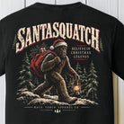 A black short-sleeve t-shirt featuring a large graphic print on the back. The graphic shows a Santa-clad Bigfoot walking through a snowy forest while carrying a lantern and a red sack, with the text 'SANTASQUATCH' and 'BELIEVE IN CHRISTMAS LEGENDS'.