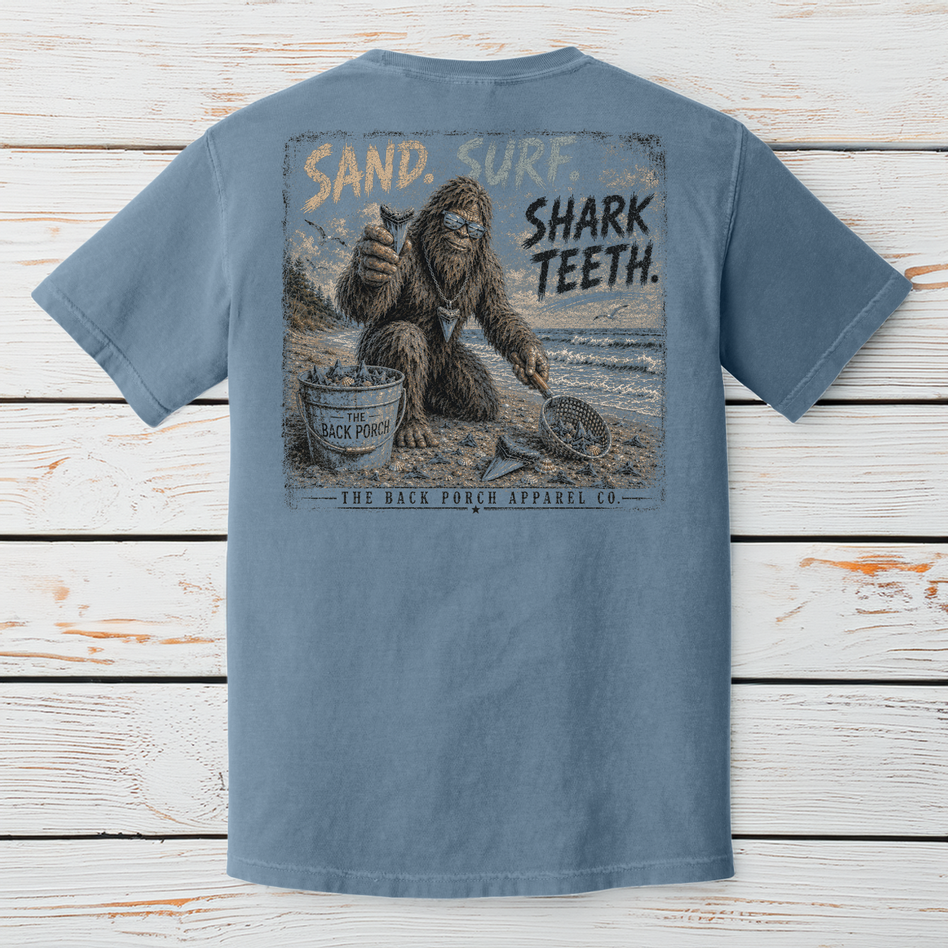 Comfort Colors Blue Jean graphic t-shirt featuring the Back Porch Apparel Co. Sand. Surf. Shark Teeth. Bigfoot beachcombing design displayed on a rustic wood background.