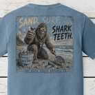 Close-up of the distressed Back Porch Apparel Co. Sand. Surf. Shark Teeth. artwork featuring Bigfoot holding a shark tooth while beachcombing with a sand scoop and bucket.