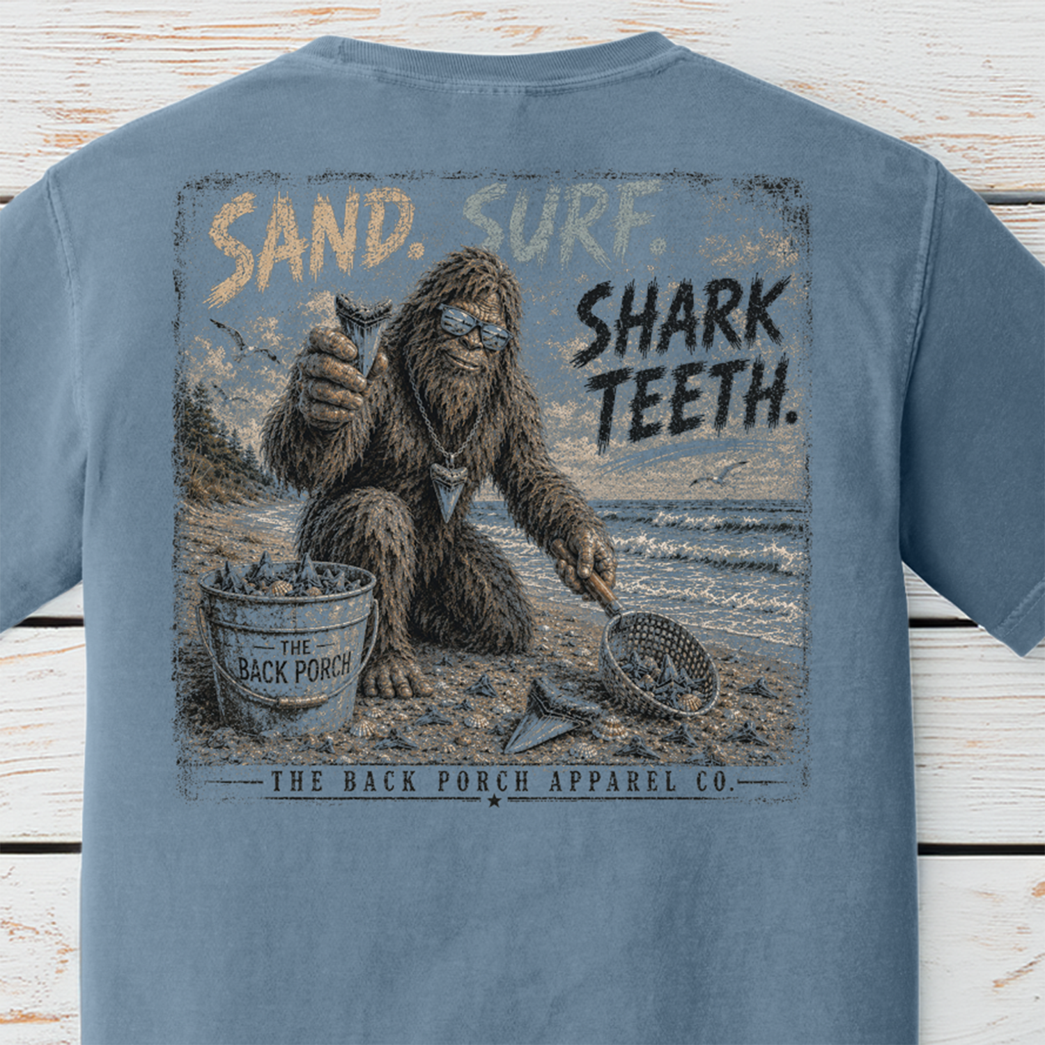 Close-up of the distressed Back Porch Apparel Co. Sand. Surf. Shark Teeth. artwork featuring Bigfoot holding a shark tooth while beachcombing with a sand scoop and bucket.