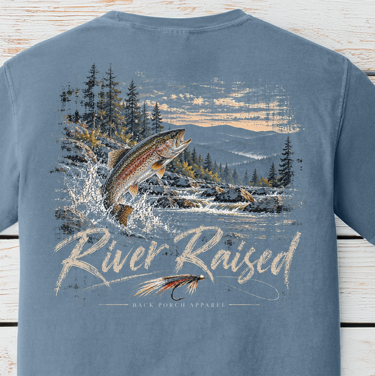 A light blue crewneck t-shirt shown from the back, featuring a large graphic print of a jumping trout in a river with mountains and pine trees, and the text 'River Raised' and 'BACK FORCH APPAREL'.