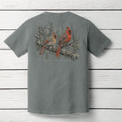 A gray short-sleeve t-shirt displayed flat against a white wood plank background, showing the back side with a large graphic print of two red cardinals perched on a snowy branch.