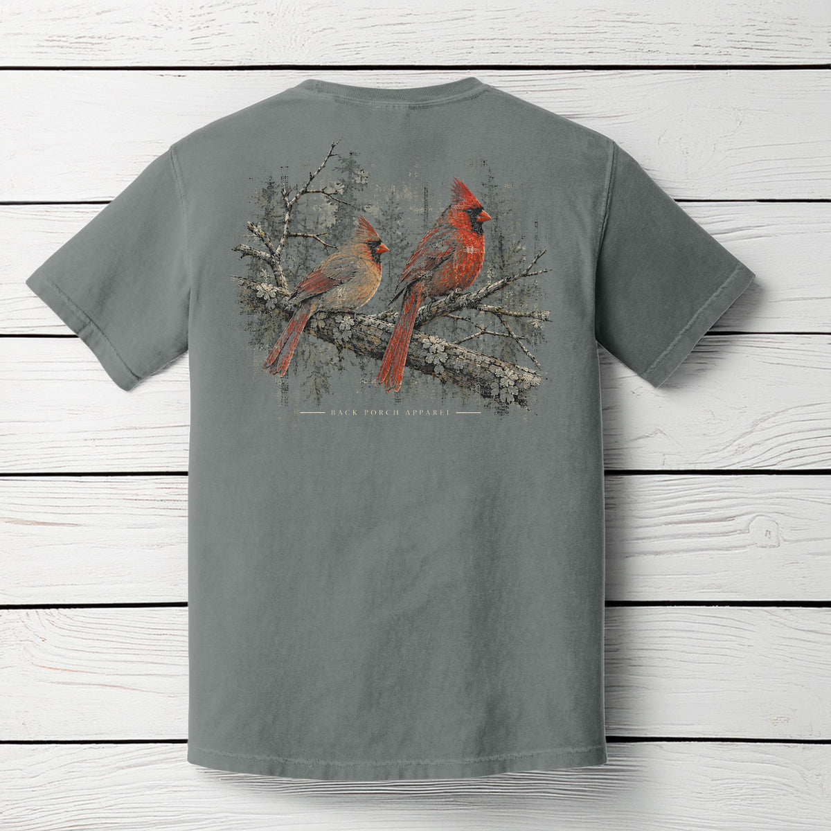 A gray short-sleeve t-shirt displayed flat against a white wood plank background, showing the back side with a large graphic print of two red cardinals perched on a snowy branch.