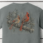 A gray short-sleeve t-shirt featuring a detailed back graphic of two cardinals perched on a branch with the text 'BACK PORCH APPAREL' underneath, displayed on a white wood plank background.