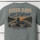 Close-up of the River Days Hit Different graphic on a Grey Comfort Colors t-shirt showing a Labrador in a fishing boat with rods, cooler, and a sunset over the river with Back Porch Apparel Co. branding.