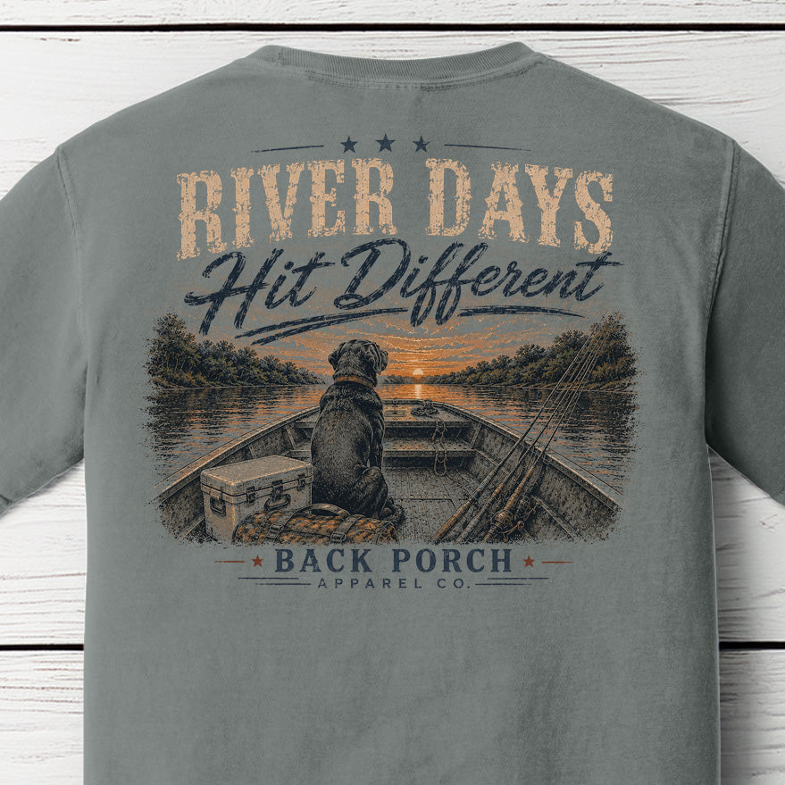 Close-up of the River Days Hit Different graphic on a Grey Comfort Colors t-shirt showing a Labrador in a fishing boat with rods, cooler, and a sunset over the river with Back Porch Apparel Co. branding.
